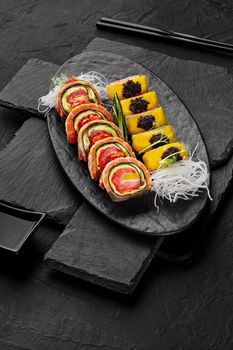 Delicious sushi rolls with tuna, salmon, ripe sweet mango and avocado slices garnished with red caviar and tobiko served on slate board with grated daikon and soy sauce on black background