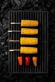 Grilled ears of corn on wooden skewers and multicolored bell peppers lying on black cast iron barbecue grate over cold coals, top view