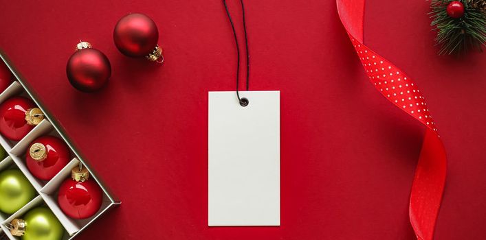 Christmas sale and holiday fashion design concept. Blank clothing tag and xmas decoration and ornaments on red paper background as flatlay mockup.