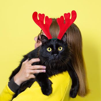 Girl in a yellow dress holds her beloved fluffy funny black cat with yellow eyes in Christmas Reindeer Antlers Headband in on a yellow background. Christmas cat