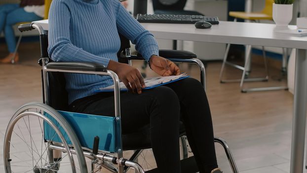 Black woman with disabilities, invalid, disabled paralized handicapped sitting immobilized in wheelchair holding clipboard with financial documents. African manager working in business office room