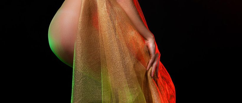 Young nude woman posing in studio with a golden shawl on a dark background. View from the back. 