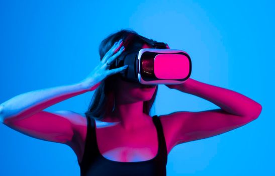 Cheerful girl with hands up wearing the virtual reality goggles and playing a games in neon light. Future technology concept. Metaverse