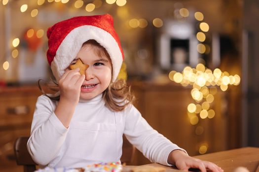 Adorable little girl in santa hat close one eye by gingerbread and laughing. Happy little kid preparing for Christmas. Background of fairy lights bokeh.