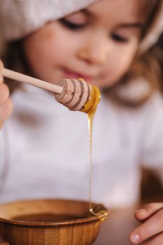 Little girl in red santa hat eating honey at kitchen decorated by fairy lights. Christmas mood at home. Happy New Year.