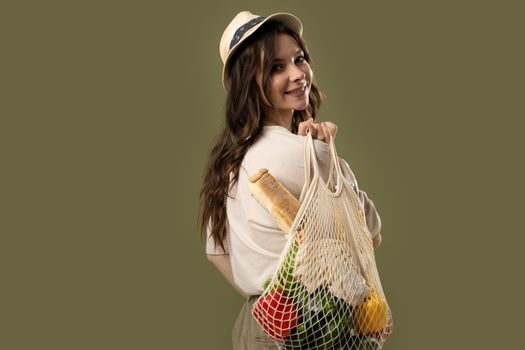 Smiling young woman in light summer clothes with a mesh eco bag full of vegetables, greens watching in a camera on a green studio background. Sustainable lifestyle. Eco friendly concept. Zero waste
