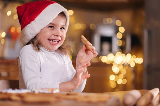 Adorable little girl in santa hat eating homemade gingerbread and laughing. Happy little kid preparing for Christmas. Background of fairy lights bokeh.