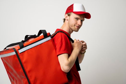 Young courier with thermal bag on color background, space for text. Food delivery service. Delivery guy in a red t-shirt uniform work as courier and holds red thermal food backpack. Service concept