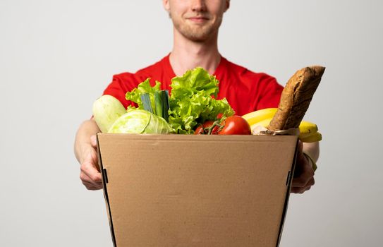 Delivery male employee in a red uniform holds paper cardbox package with groceries. Products delivery from shop or restaurant to home. Copy space