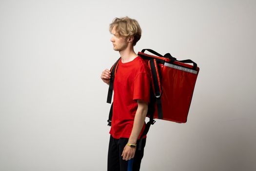 Portrait of a young courier, delivery man in red uniform with a thermal backpack isolated on a white background. Fast home delivery. Online order. Courier delivers groceries home