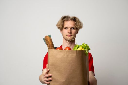 Food delivery service. Portrait of pleased delivery man in red uniform smiling while carrying paper bag with food products isolated over white background