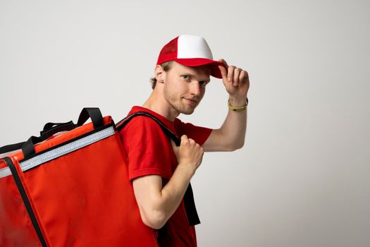 Young courier with thermal bag on color background, space for text. Food delivery service. Delivery guy in a red t-shirt uniform work as courier and holds red thermal food backpack. Service concept