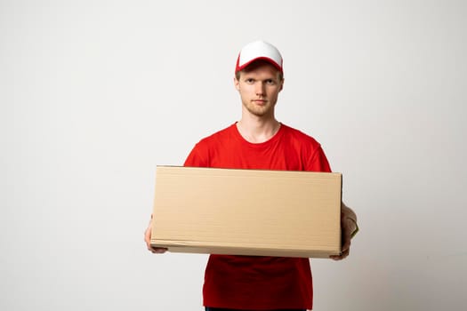 Delivery service. Young smiling courier holding cardboard box. Happy young delivery man in cap and red t-shirt standing with parcel isolated on white background