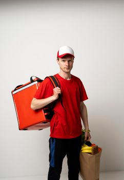 Delivery man in red uniform with a thermal bag on a shoulder holding paper bag with groceries. Food delivery service
