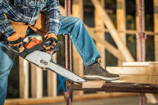 Construction Contractor Cutting Wood Board Using Powerful Chainsaw Power Tool. Wooden Skeleton Structure of Residential Building. Construction Industry Theme.