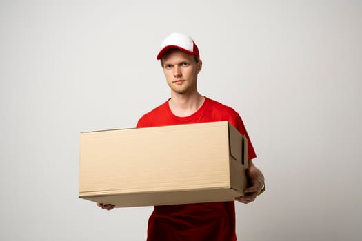 Delivery service. Young smiling courier holding cardboard box. Happy young delivery man in cap and red t-shirt standing with parcel isolated on white background