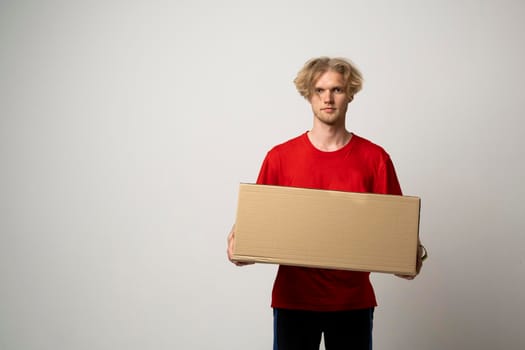 Delivery service. Young smiling courier holding cardboard box. Happy young delivery man in cap and red t-shirt standing with parcel isolated on white background