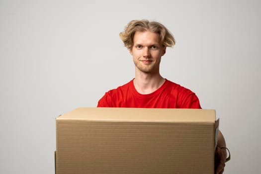 Delivery service. Young smiling courier holding cardboard box. Happy young delivery man in cap and red t-shirt standing with parcel isolated on white background