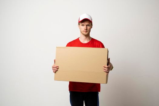 Male employee in in red uniform working as courier dealer and holds cardboard box isolated on white background, studio portrait. Service concept. Mock up copy space