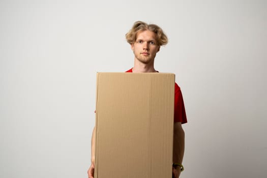 Delivery service. Young smiling courier holding cardboard box. Happy young delivery man in cap and red t-shirt standing with parcel isolated on white background