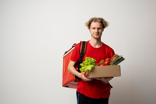 Happy courier man in t-shirt and thermal bag backpack holds paper box with food products isolated on red background studio. Delivery service from groceries market to home