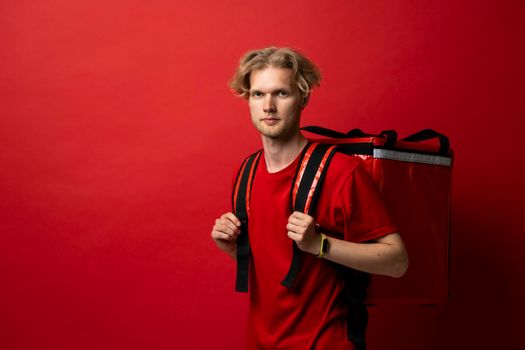 Portrait of a young courier, delivery man in red uniform with a thermal backpack isolated on a red background. Fast home delivery. Online order. Courier delivers groceries home