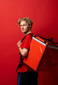 Young courier in a red uniform t-shirt and with red food thermo bag on a shoulder standing isolated on red background studio. Food delivery service