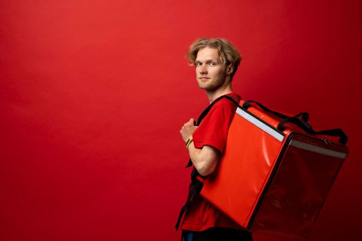 Young courier with thermal bag on red background, space for text. Food delivery service. Delivery guy in a red t-shirt uniform work as courier and holds red thermal food backpack. Service concept