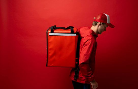 Side view of young courier in a red uniform and with a thermo bag on color red background, space for text. Food delivery service