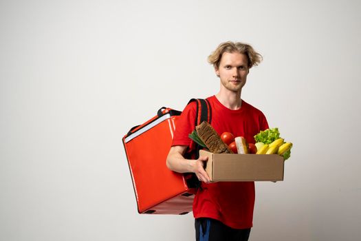 Food delivery service. Portrait of pleased delivery man in red uniform smiling while carrying paper box with food products isolated over white background