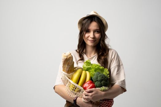 Smiling young woman in light summer clothes with a mesh eco bag full of vegetables, greens watching in a camera on a green studio background. Sustainable lifestyle. Eco friendly concept. Zero waste