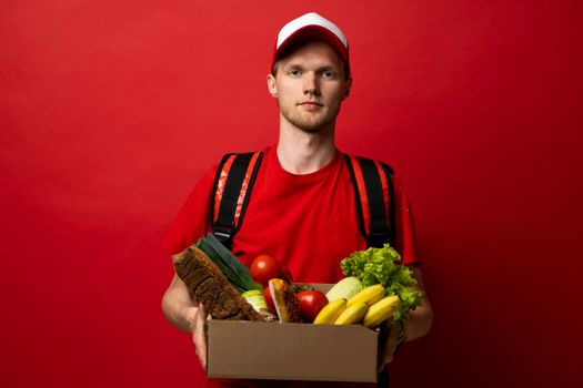 Delivery Concept. Portrait of pleased delivery man in red uniform smiling while carrying paper bag with food products isolated over red background. Food delivery service