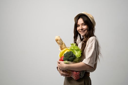 Portrait of happy smiling young woman in beige oversize t-shirt holding reusable string bag with groceries over white background. Sustainability, eco living and people concept