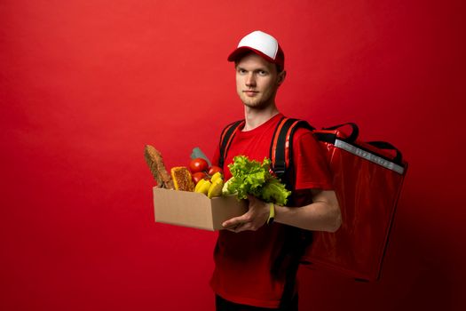 Delivery male employee in a red uniform with a thermal bag holds paper cardbox package with groceries on a red background. Products delivery from shop or restaurant to home. Copy space