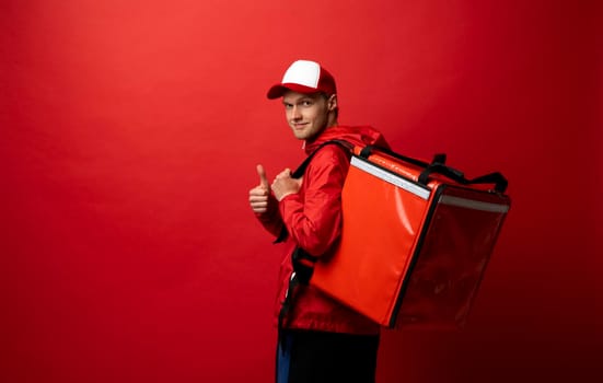 Delivery guy employee man in red cap and uniform workwear work as dealer courier hold red thermal food bag and showing thumb up isolated on red color background studio. Service concept