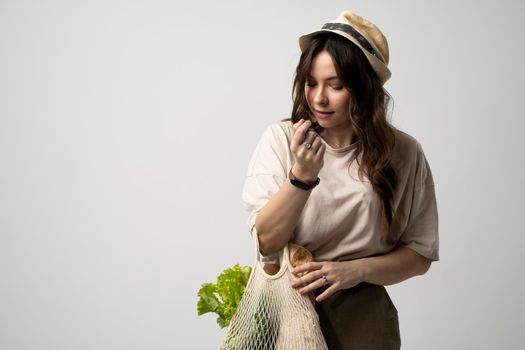 Happy woman in a beige t-shirt and a hat holding reusable cotton shopping bag with groceries, bread and greens. Concept of no plastic. Zero waste, plastic free. Sustainable lifestyle