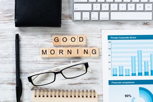 Good morning message with letters on wooden cubes. Still life of workspace with supplies. Flat lay wooden desk with computer keyboard and spiral notebook. Morning greeting phrase and motivation.