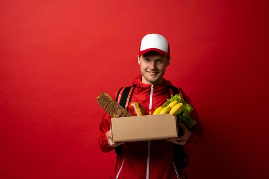 Portrait of smiling handsome delivery man holding paper grocery box with a groceries isolated on red background