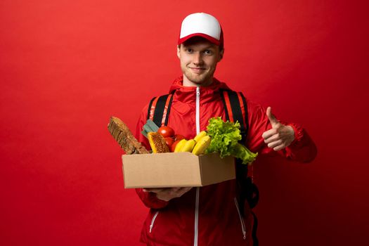 Grocery delivery courier in red uniform with a paper box with food showing a thumb up. Thumb up gesture. Food delivery service concept