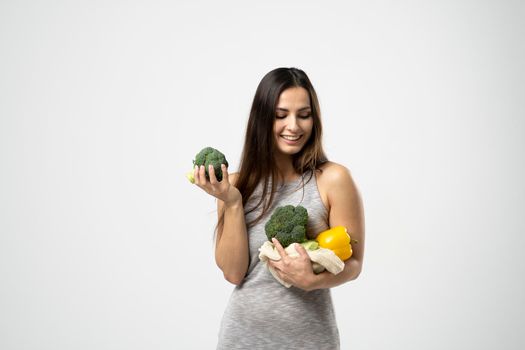 Young brunette girl holds vegetables in mesh eco bag in one hand and a broccoli in another. Zero waste concept, plastic-free, eco-friendly shopping, vegan