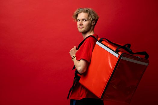 Delivery guy employee man in red cap and t-shirt uniform workwear work as dealer courier hold red thermal food bag backpack isolated on red color background studio. Service concept