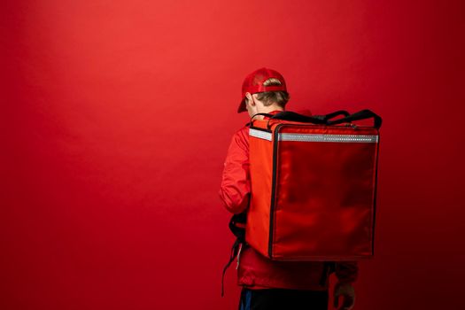 Young courier in a red uniform t-shirt and with red food thermo bag on a shoulder standing isolated on red background studio. Food delivery service