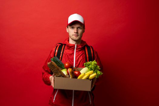 Young delivery man carrying paper box bag of grocery food and drink from store isolated on red studio background. Copy Space. Delivery Concept