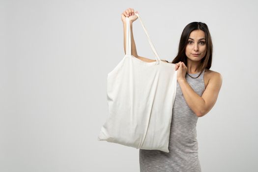 Cheerful millennial woman showing white eco bag to camera standing over white studio background. Lady holding cotton shopper handbag. fashion and ecology concept