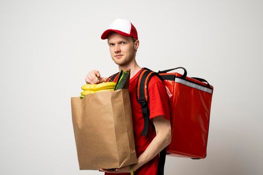 Delivery Concept - Handsome delivery man in a red uniform with thermal backpack carrying paper package bag of grocery food and drink from grocery store. Isolated on white background. Copy Space