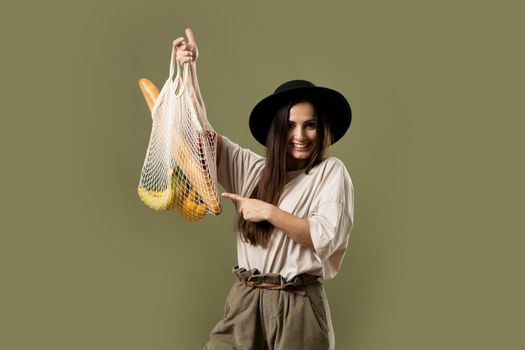 Mesh eco bag with groceries, vegetables and fruits, in the hands of a brunette girl in a beige t-shirt and black hat. Eco friendly concept. Woman holding string shopping bag