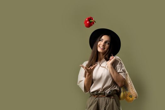 Smiling brunette young woman in a beige t-shirt and a hat holding a reusable mesh bag with fresh vegetables and fruits on a shoulder and throwing a red pepper in a air. Zero waste concept