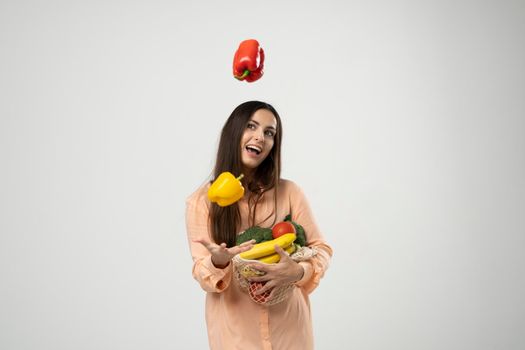 Smiling woman in dress throw up a red and yellow pepper in a air by one hand and holding reusable cotton eco bags in other