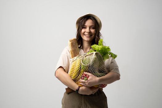 Eco friendly positive young woman in beige oversize t-shirt holding reusable mesh cotton eco bags for shopping with groceries on white background