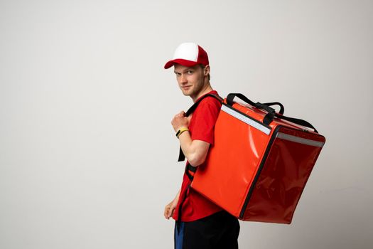 Young courier in a red uniform t-shirt and with red food thermo bag on a shoulder standing isolated on white background studio. Food delivery service